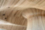Curved wooden structure with vertical stripes forms a fluid, wave-like pattern. Light wood tones dominate the abstract architectural design.