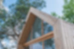 Wooden frame cabin with large windows against a clear blue sky and tall green trees, evoking a peaceful, natural setting.